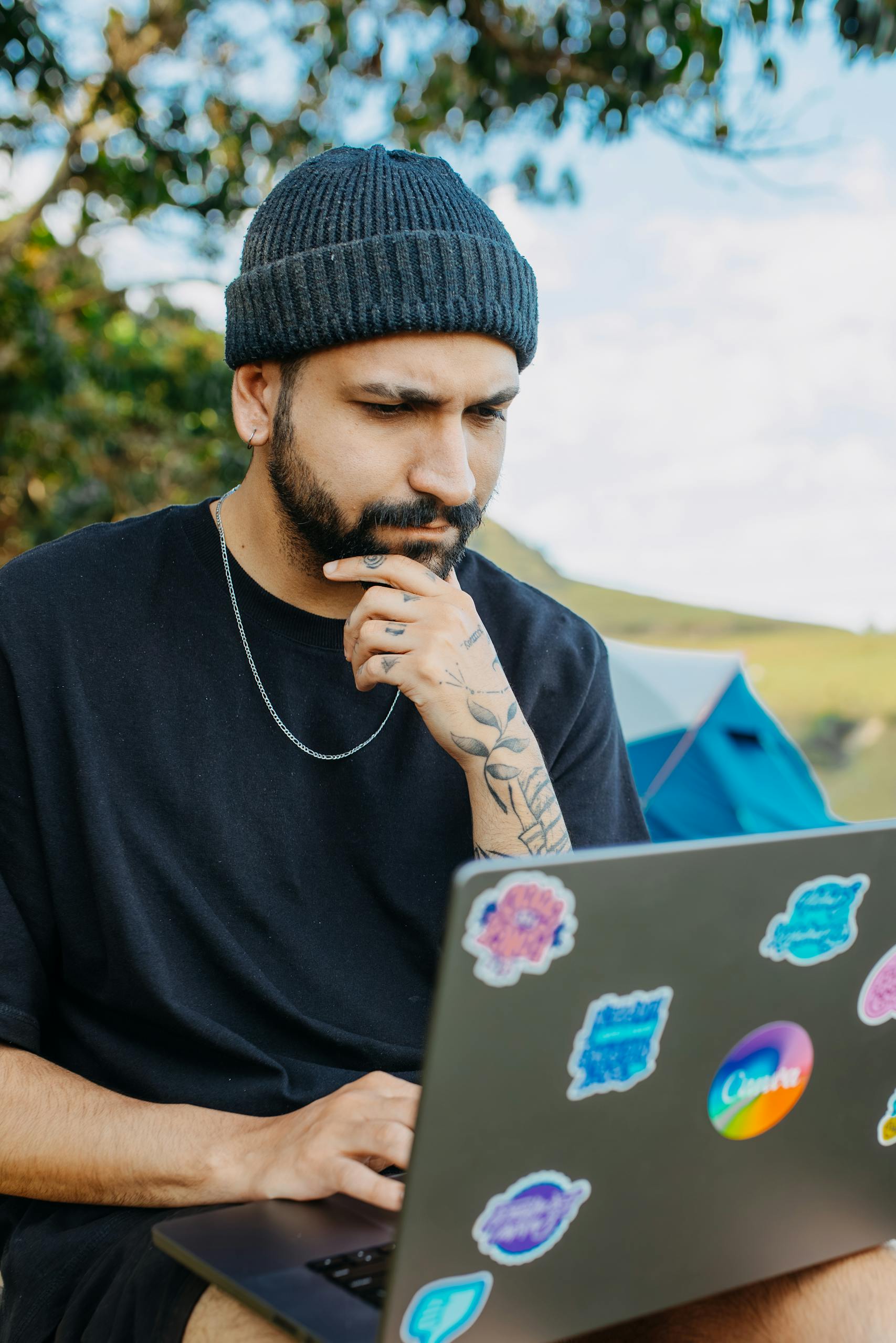 Young man with tattoos sitting outdoors, using a laptop while camping and surrounded by nature.